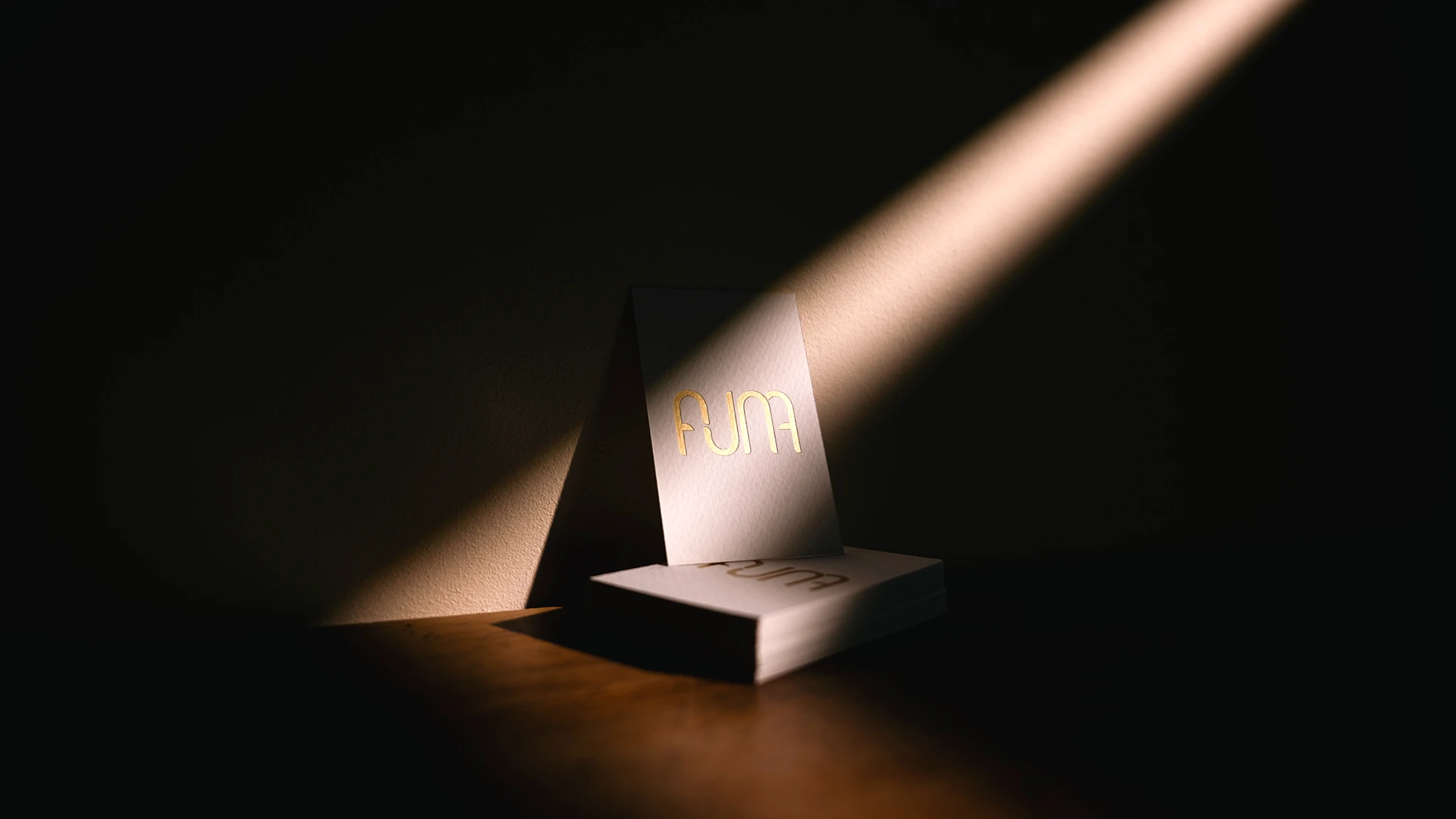 Carte de visite Studio Auna – photographie avec dorure à chaud Photographie en clair-obscur d'une pile de cartes de visite Studio Auna avec logo doré à chaud, éclairées par un rai de lumière sur fond noir — mise en page et identité visuelle par Heji